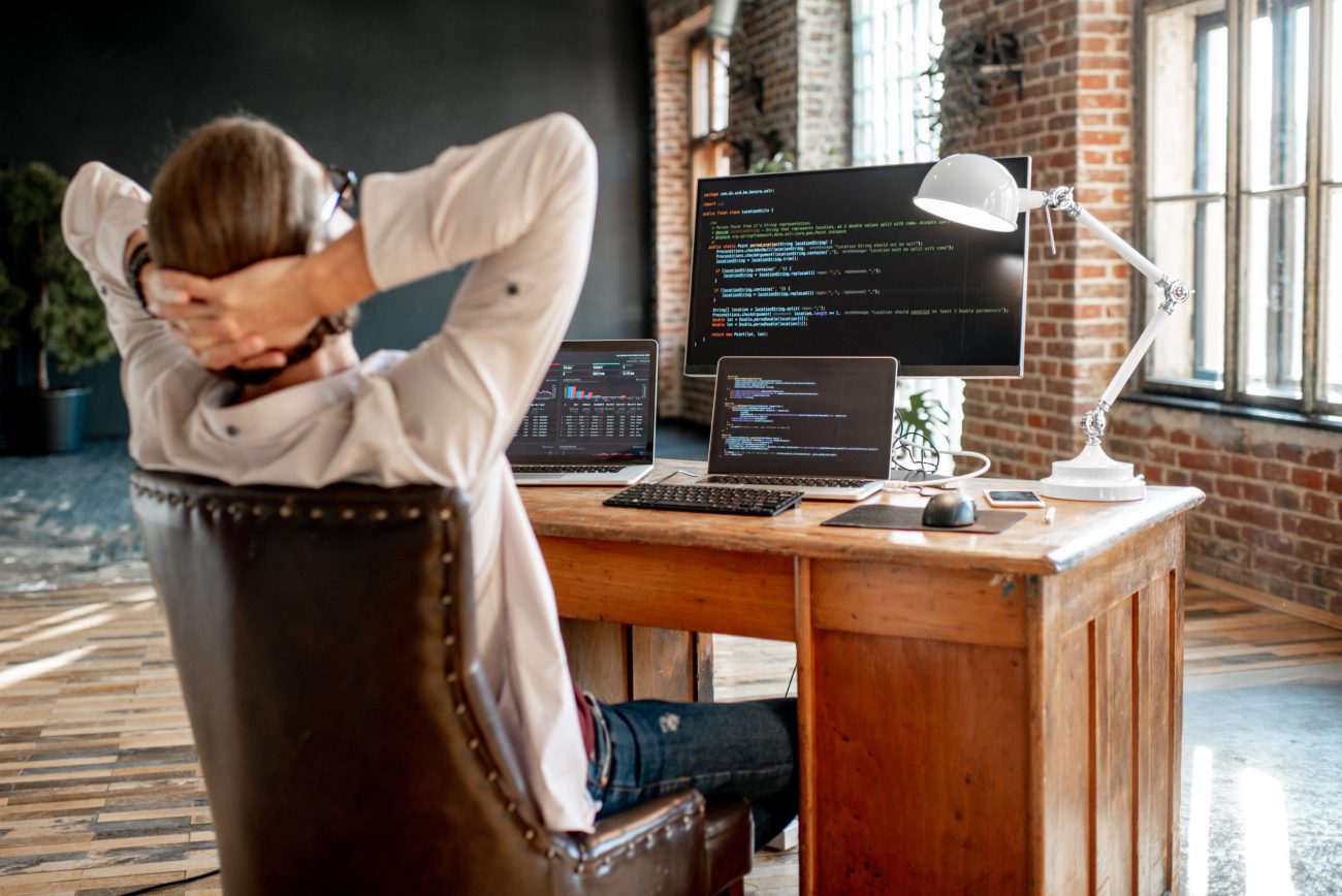 Young male programmer relaxing during the work with program code sitting at the workplace with three monitors in the office. Image focused on the screen